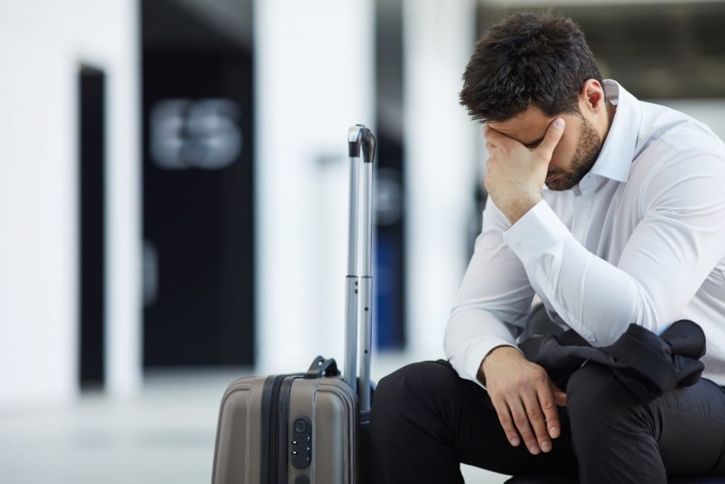 Homem triste em aeroporto com sua bagagem após ter o embarque negado por overbooking.